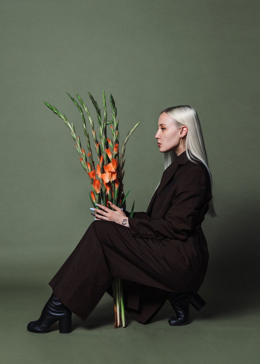 Jessica De La Pair in profile, seated in a brown suit holding orange gladioli against an olive backdrop